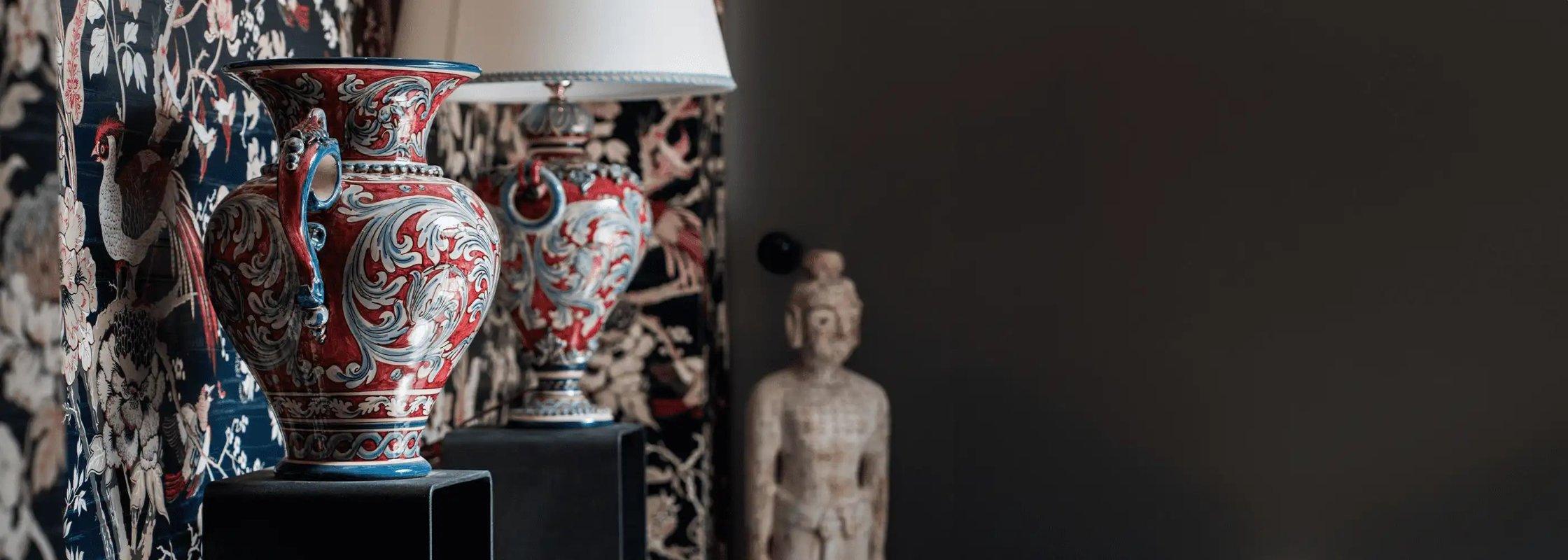 Ceramiche Caltagirone fatti a mano, vasi decorativi rossi e blu con motivi classici su supporti neri, lampada e statua sfocata