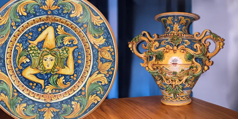 Un Vaso modellato e decorato a mano con un figurato veliero, sulla sinistra un piatto con una raffigurazione di una trinacria in colorazione Blu, Arancio e Verde