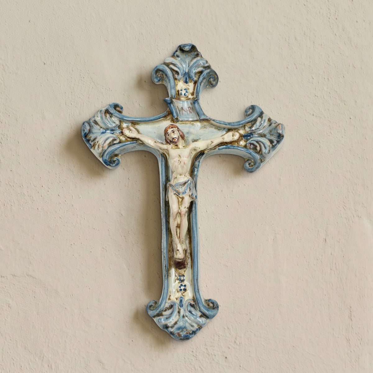 Mono Blue Caltagirone ceramic crucifix hanging on a light wall, handmade with Christ in relief and hand-painted blue scrolls